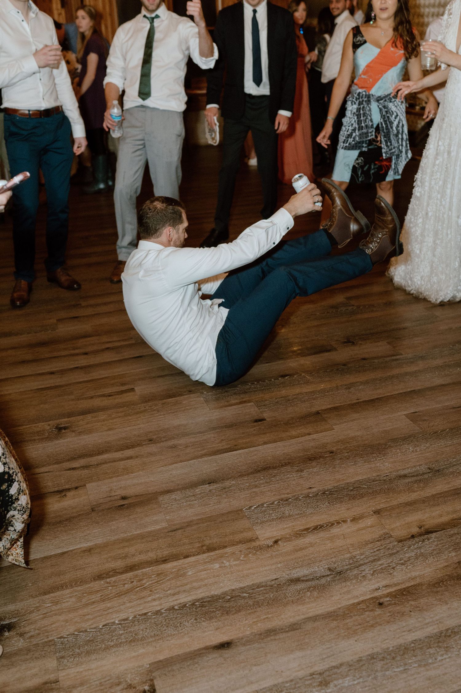 Wedding guest performs dramatic dance move on wooden floor during reception celebration.