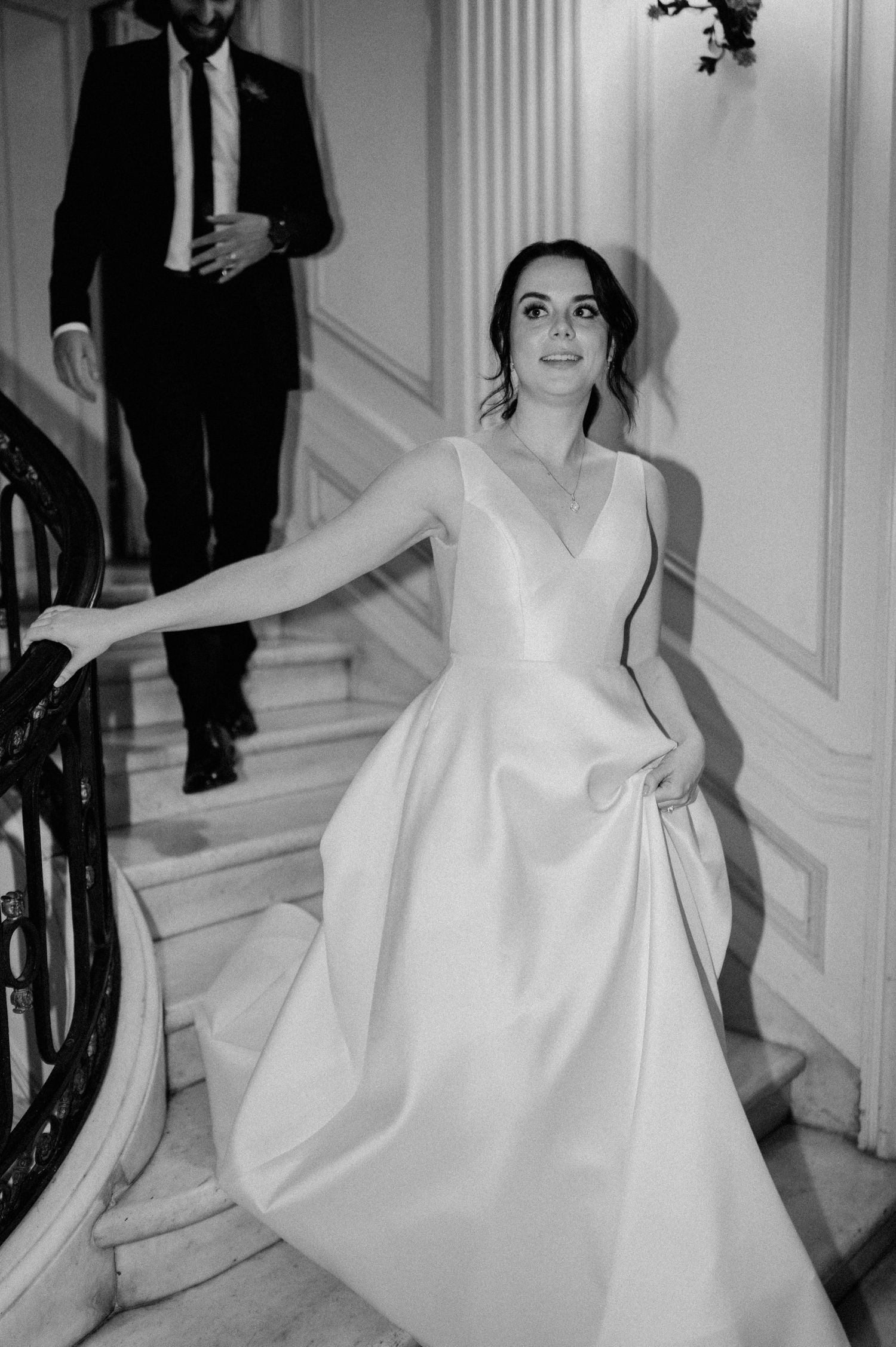 Bride in elegant white wedding gown poses on classic staircase with white molded walls.