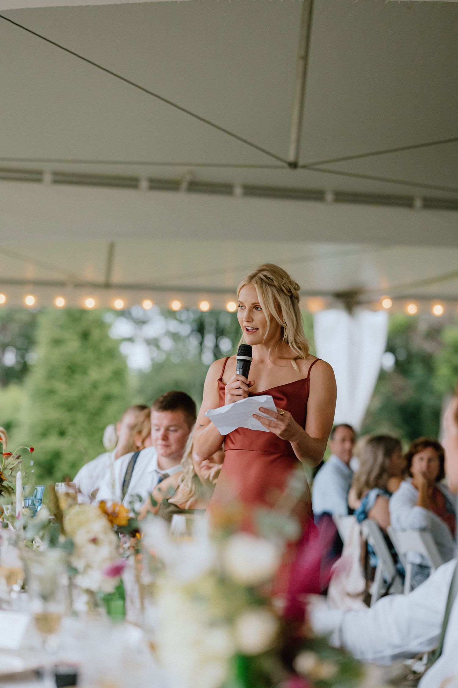 Wedding guest in burgundy dress giving speech during outdoor tent reception with glowing string lights.