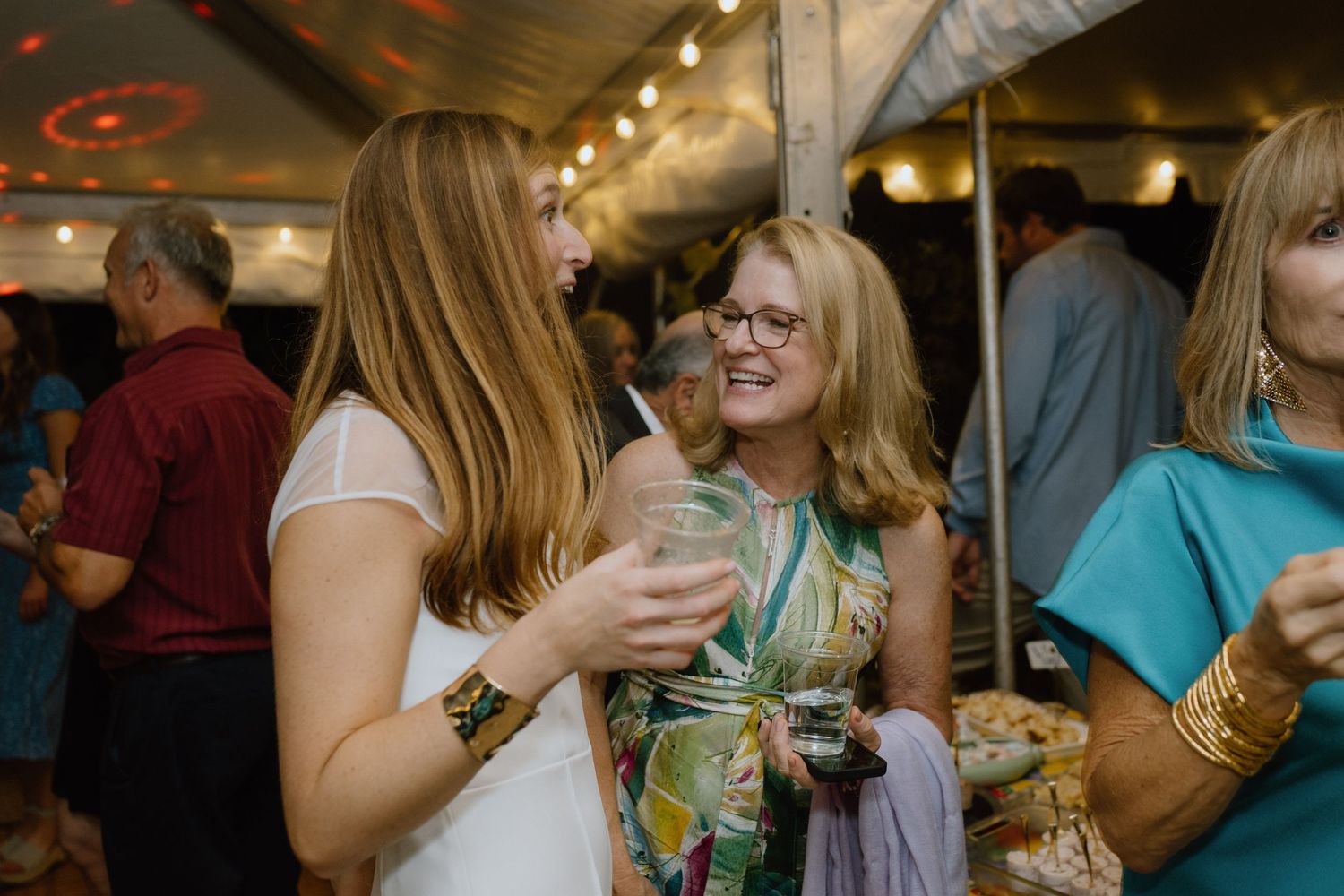 Party guests laughing and enjoying drinks at a celebration under a tent.
