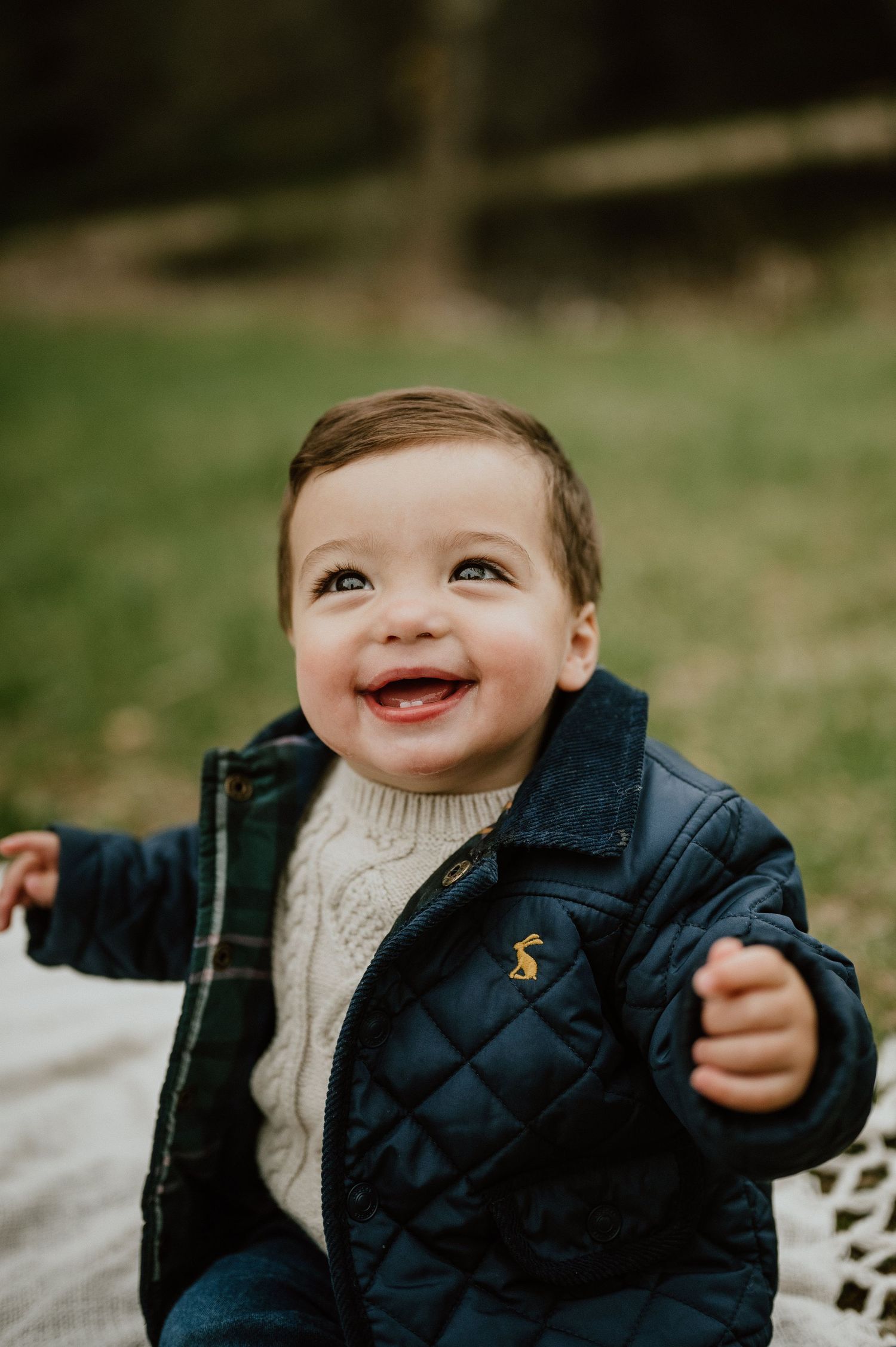 Toddler wearing navy quilted jacket and cream sweater smiles joyfully outdoors.