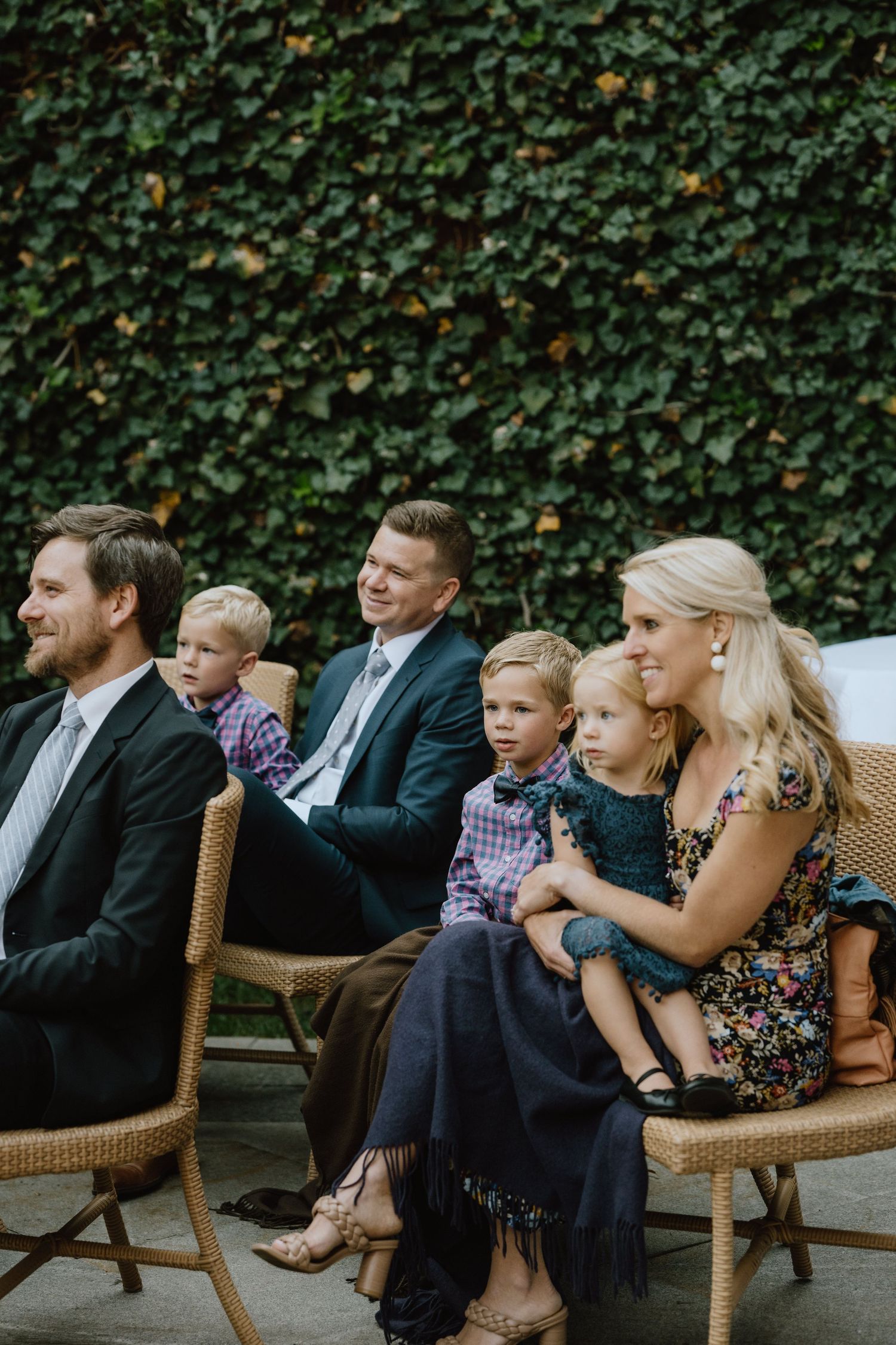 Wedding guests seated in wicker chairs against a lush ivy wall backdrop during an outdoor ceremony.