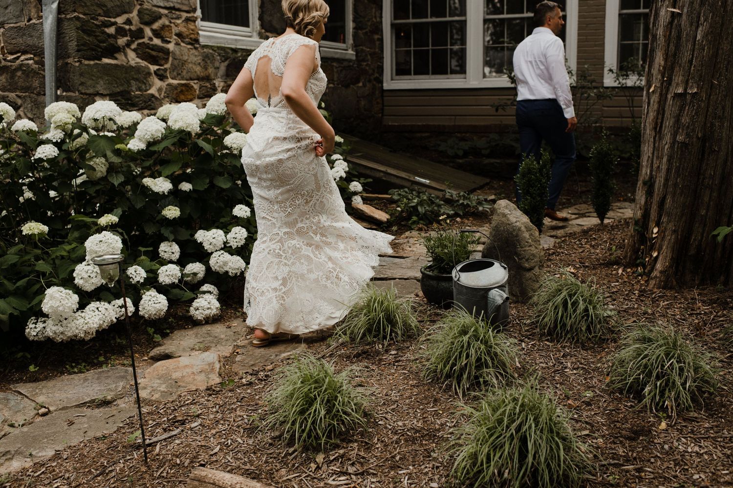 A bride in a lace gown walks through a garden with blooming white hydrangeas.