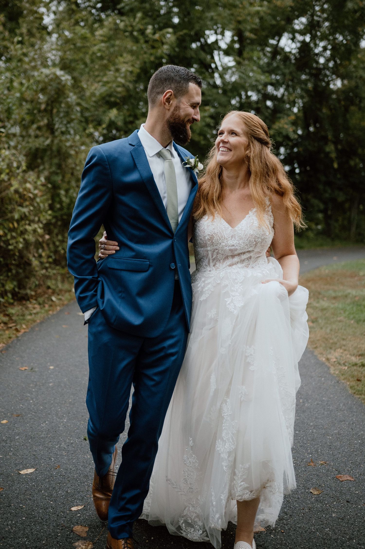 A couple in wedding attire walk together and laugh on a paved path surrounded by trees.