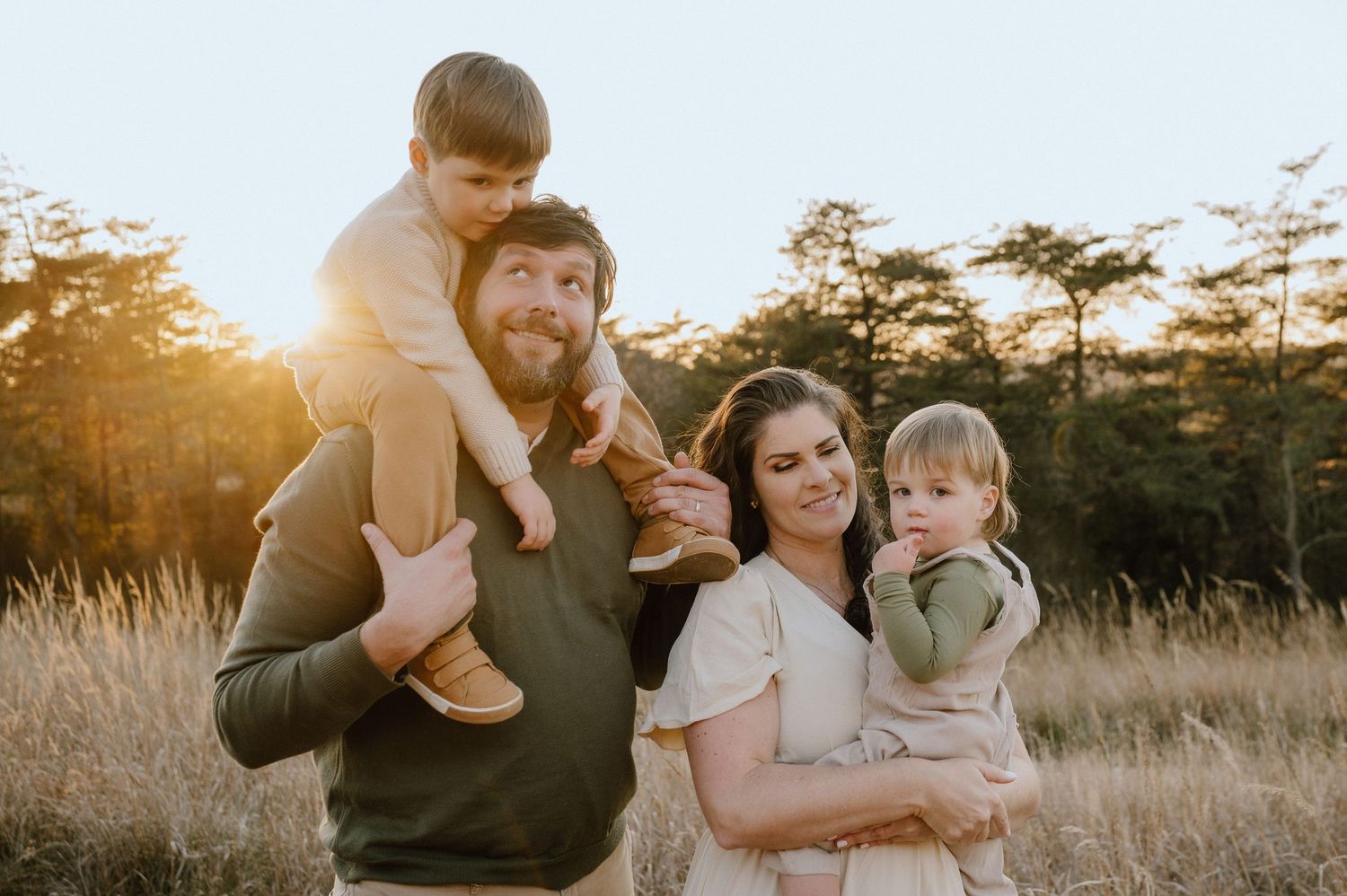 Family portrait captured during golden hour in a field with evergreen trees in the background.