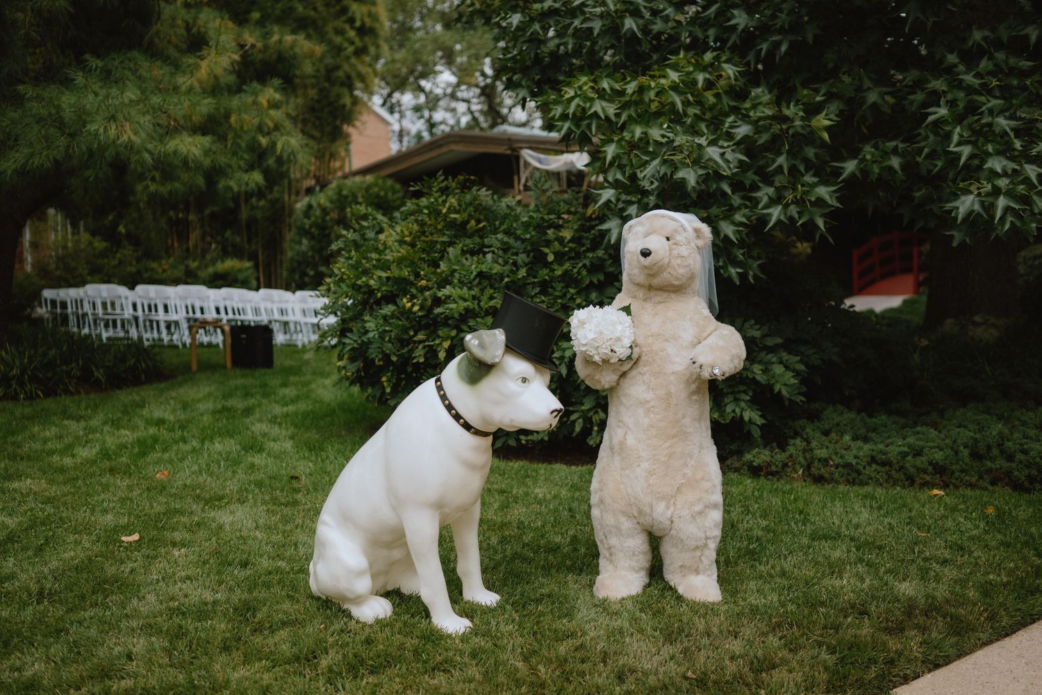 White dog sitting next to large plush teddy bear on grass with chairs in background.
