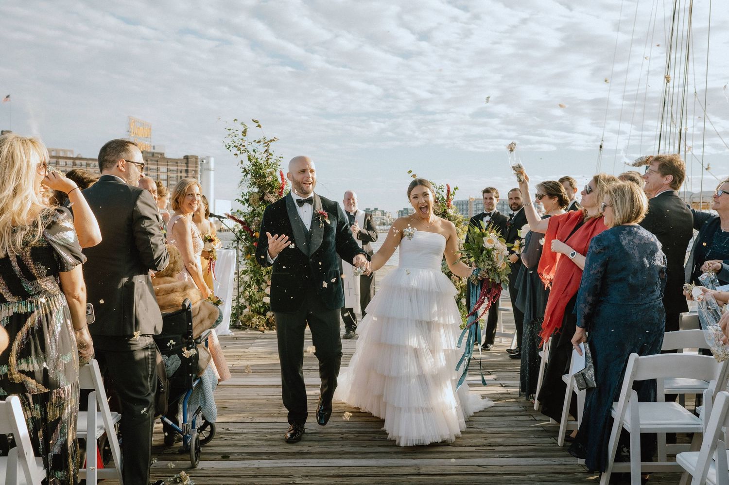 Bride and groom walk down outdoor wedding aisle on waterfront dock, guests cheering and tossing confetti.