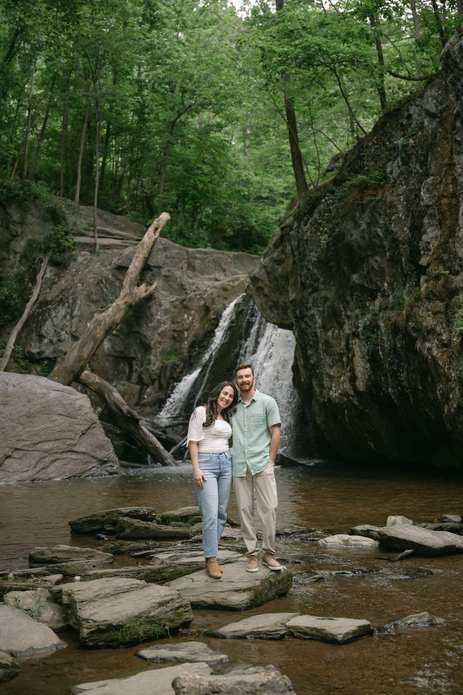 Happy couple standing on stepping stones in front of a waterfall in a rocky forest.