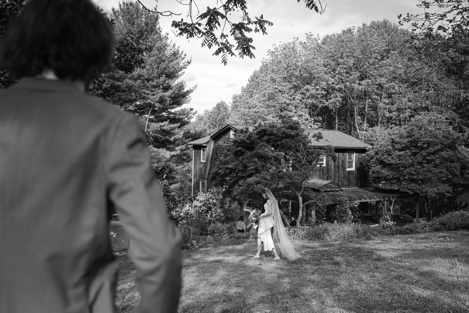 Black and white wedding photo of bride in gown near rustic house surrounded by lush trees, with guest in foreground.