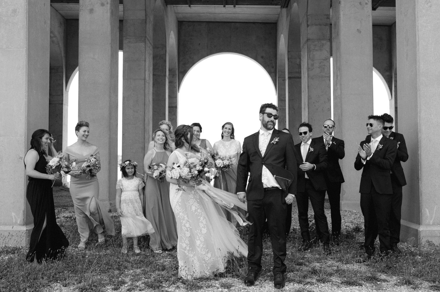 Wedding party poses together under a covered outdoor colonnade.
