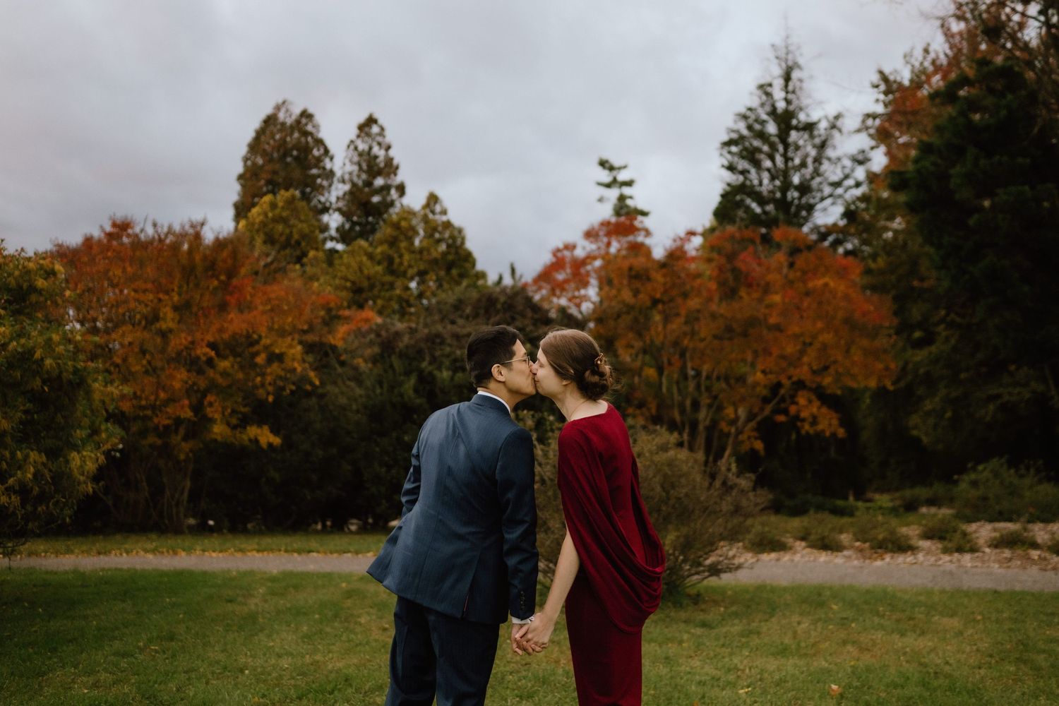 Couple sharing a kiss in a lush autumn park, man in blue suit and woman in burgundy dress, colorful fall foliage backdrop.