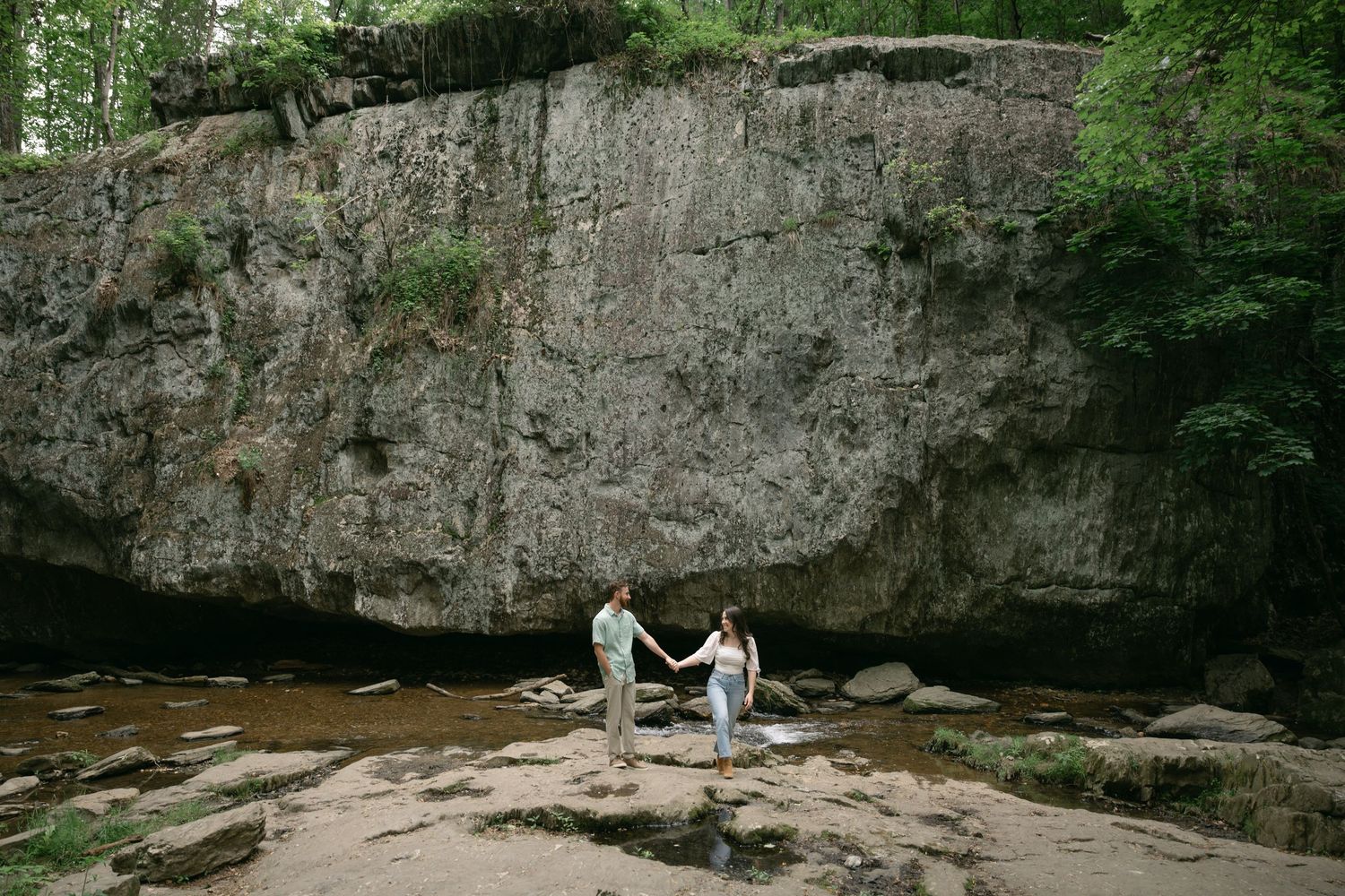 Couple holding hands on rocky riverbank before massive stone cliff wall surrounded by lush forest trees.