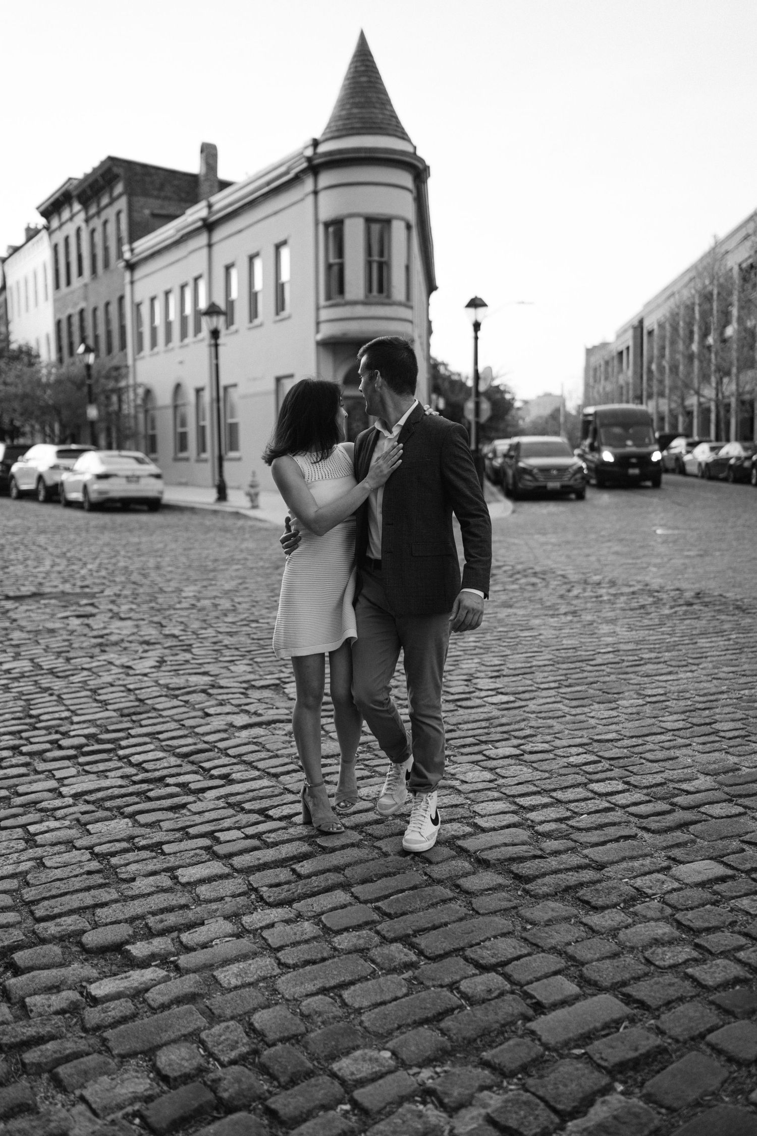 Black and white photo of a couple sharing a romantic moment on a cobblestone street with historic buildings in the background.