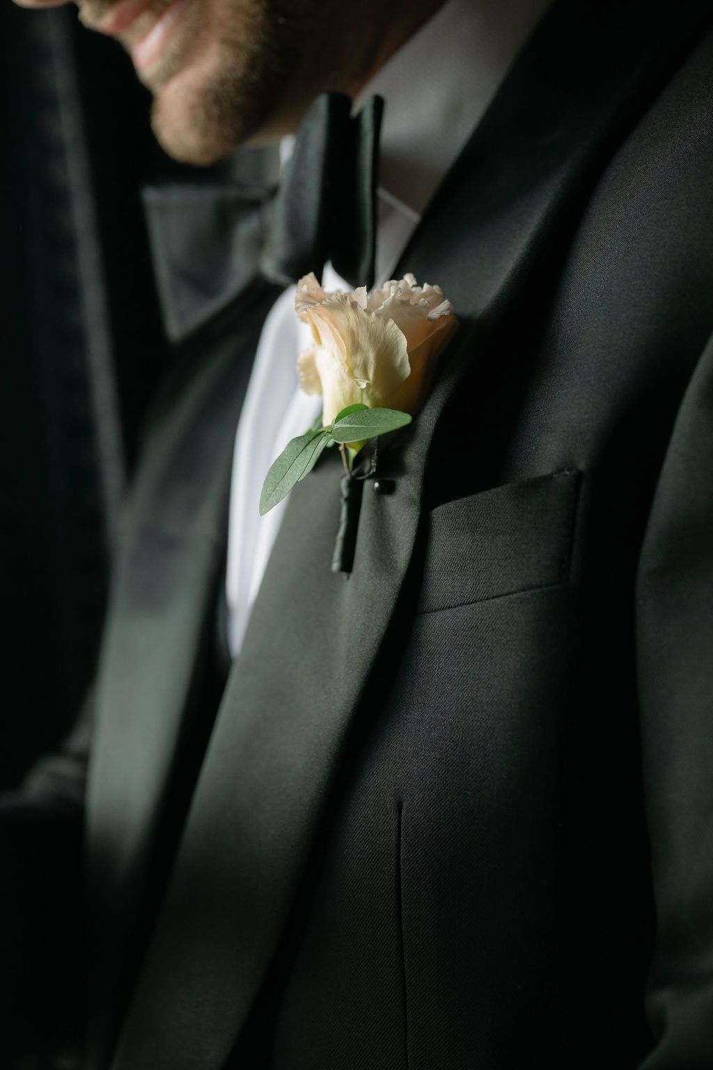 A white rose boutonniere pinned to a black formal suit lapel in an elegant wedding detail shot.