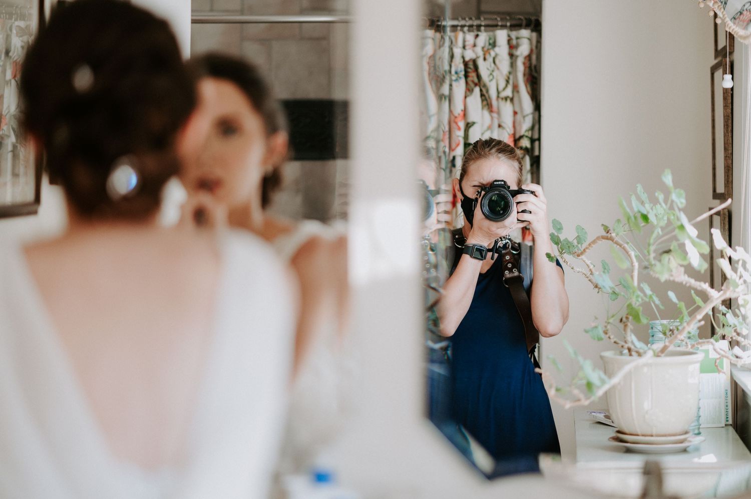 Photographer in navy dress captures mirror selfie while bride in white dress prepares in elegant bathroom.