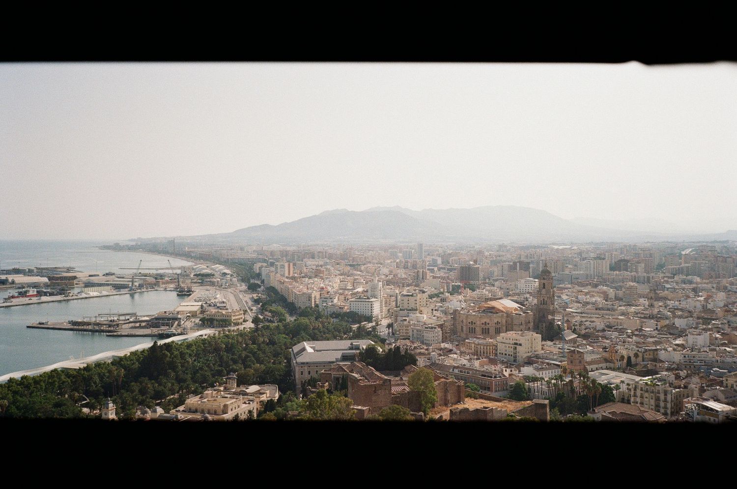 Wide panoramic view of a coastal Mediterranean city with port and mountains in background.