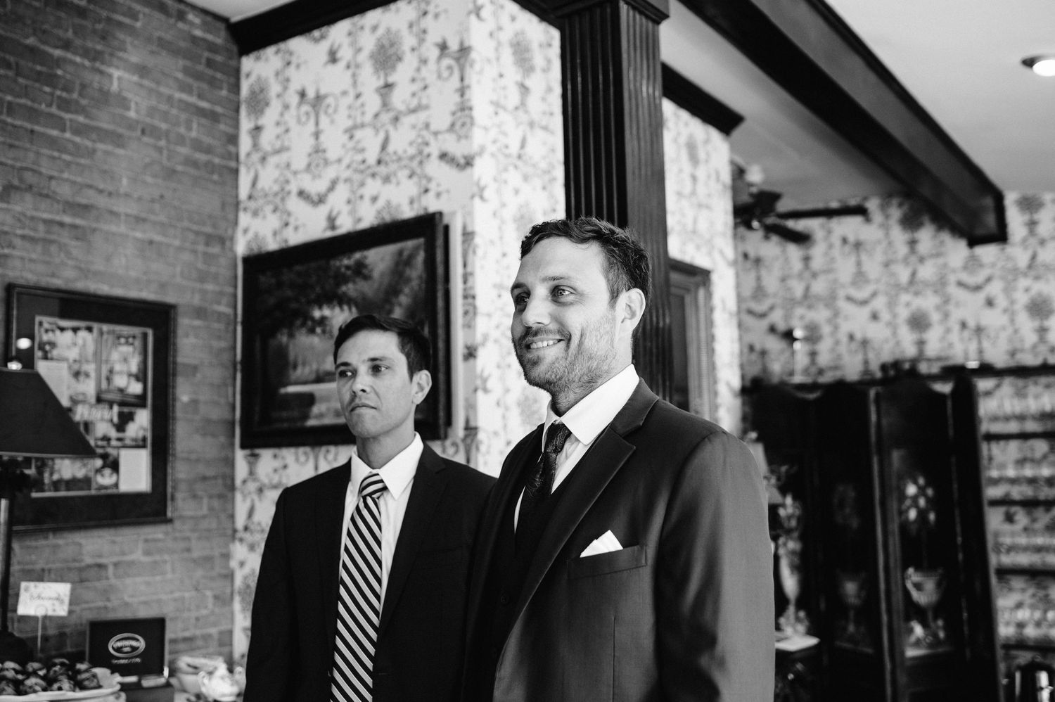 Two men in formal suits standing in brick-walled venue space.