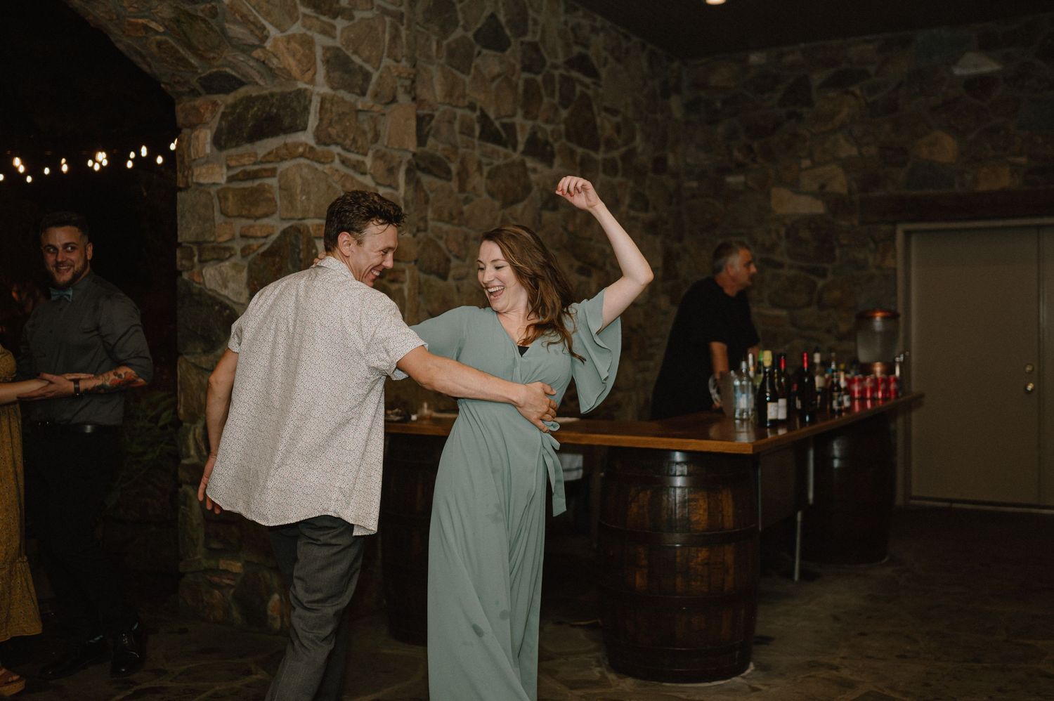 A couple dances together in a rustic stone-walled venue with wooden barrels in the background.