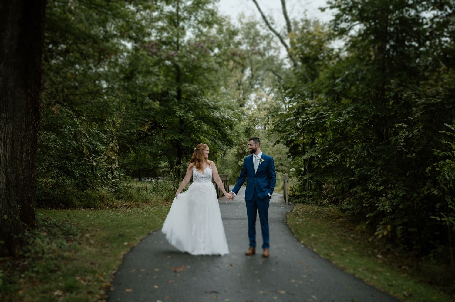 A couple in wedding attire walking together on a tree-lined path.