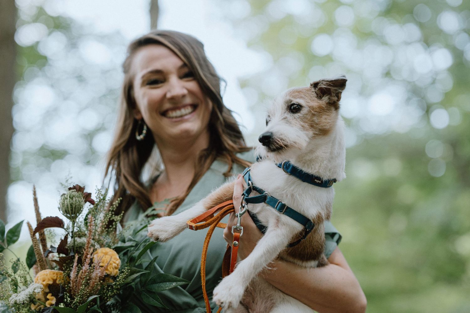 A joyful moment with a small white dog and wedding bouquet in a sunlit forest setting.