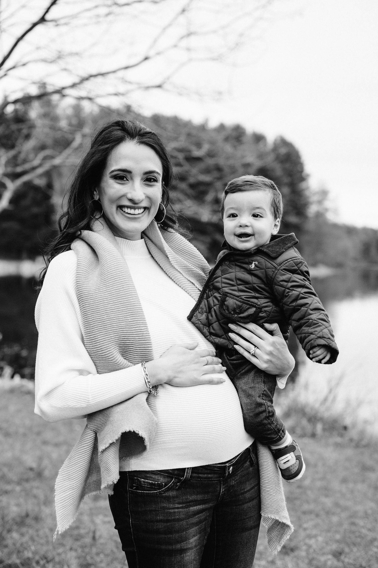 Black and white portrait of expectant parent holding toddler by lake.