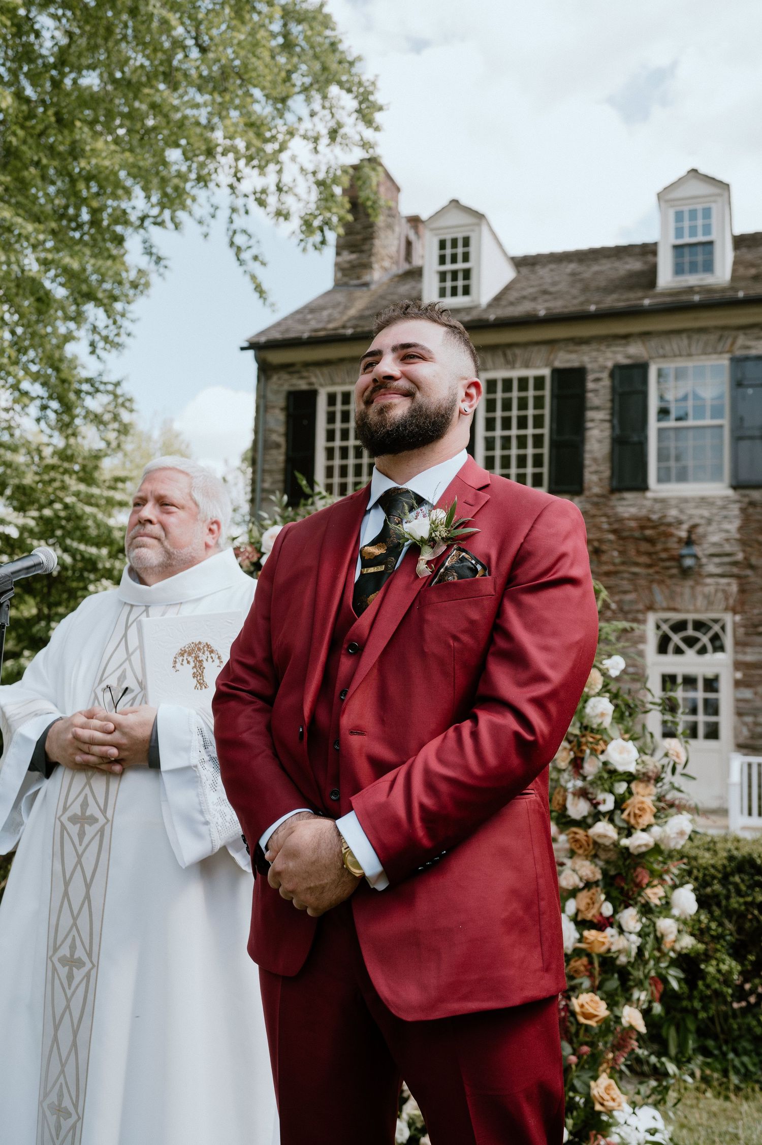 A wedding ceremony scene outside a historic stone house with clergy and groom in burgundy suit.