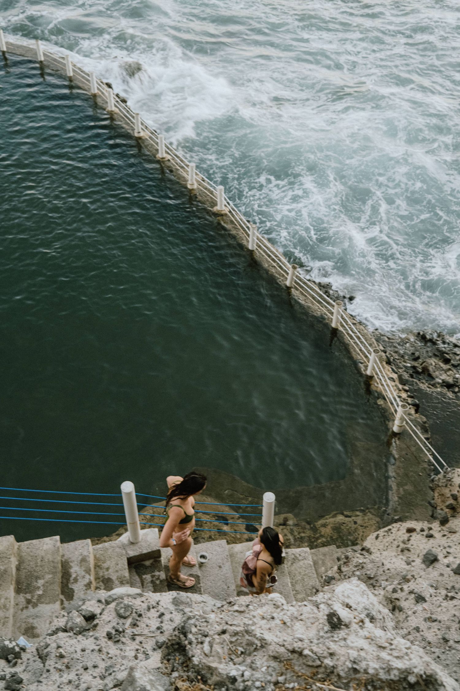 Two swimmers descend stone steps leading to a curved ocean pool with white barriers.