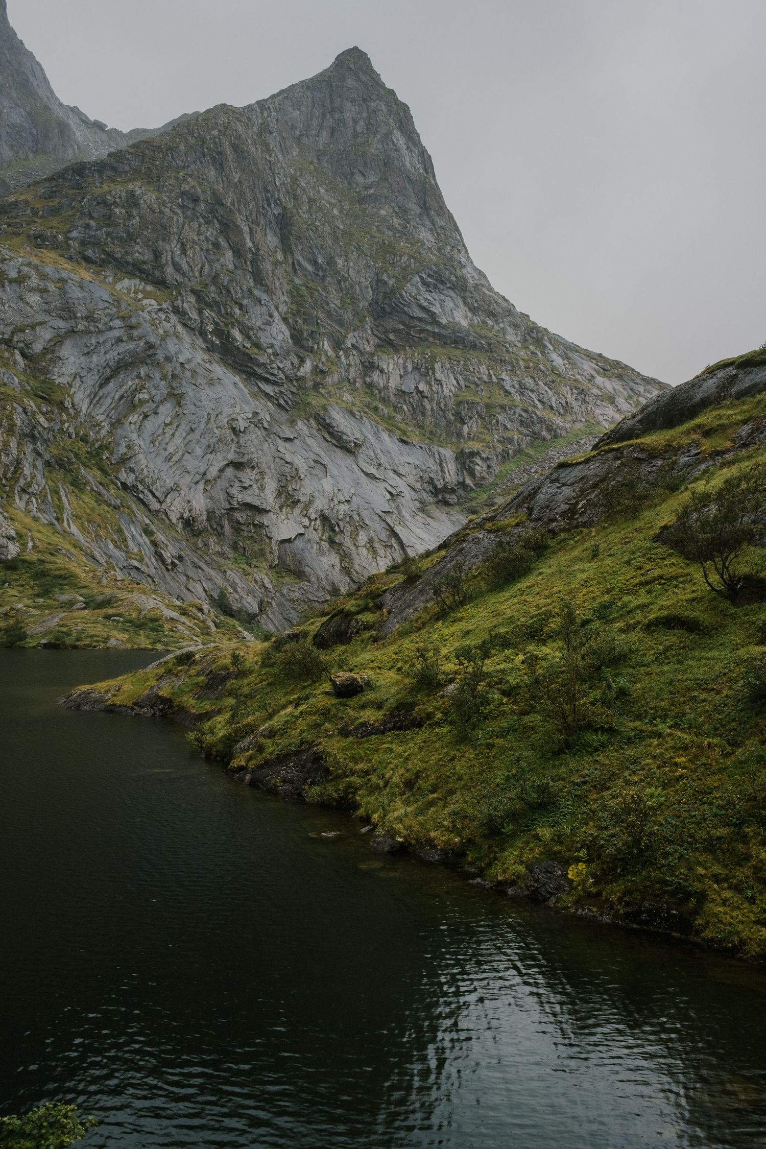 Steep mountain cliff face rising above a dark alpine lake with green slopes in foggy conditions.