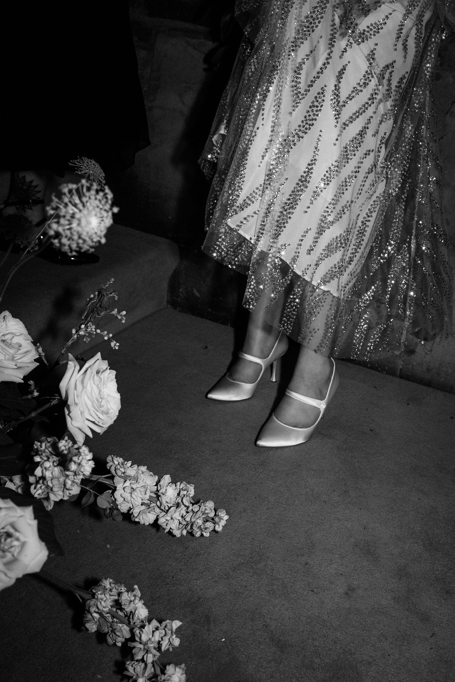 A close-up black and white photo of sparkly dress fabric and vintage shoes next to scattered flowers on the ground.