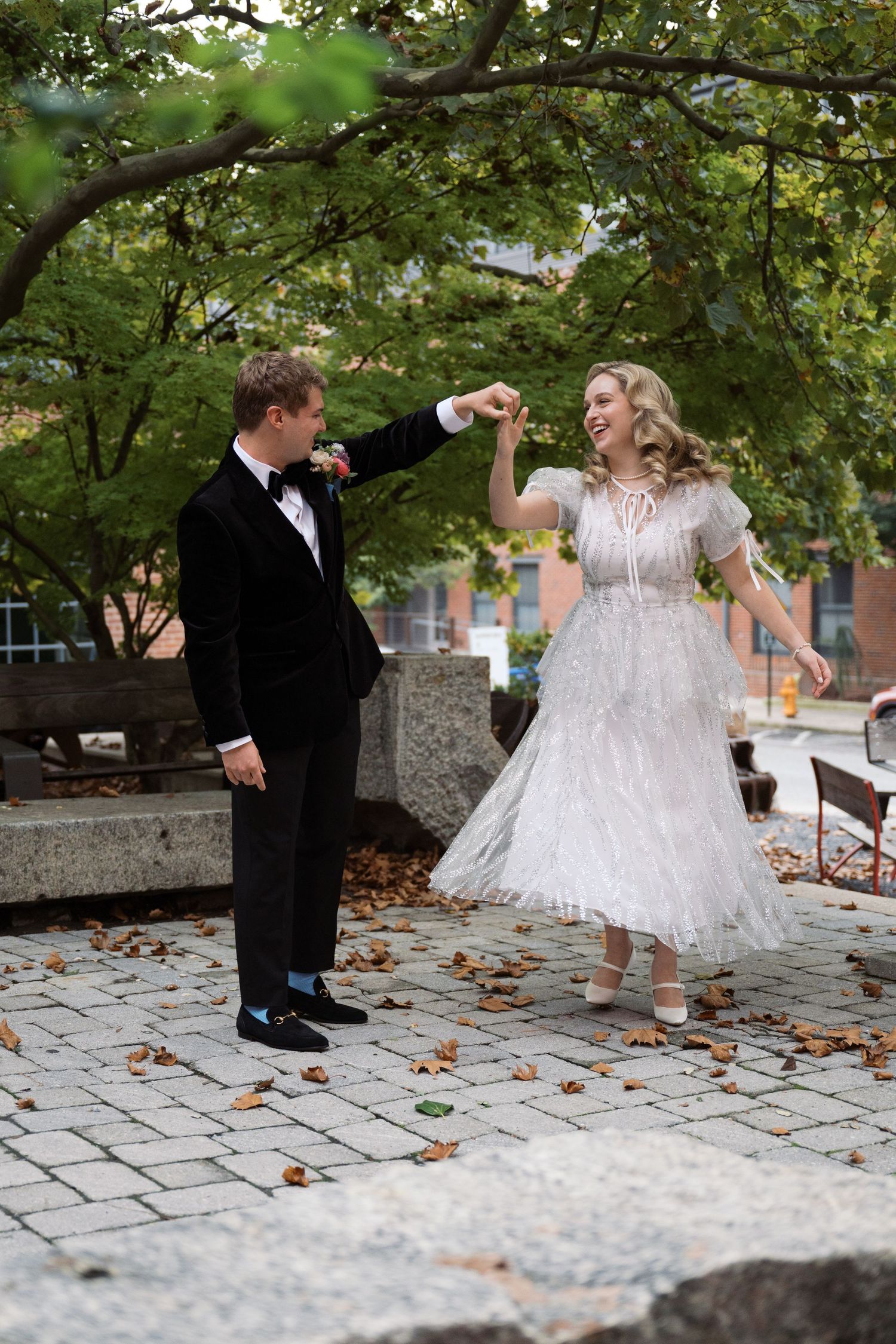 A couple dances playfully on stone pavement under green trees during their wedding celebration.
