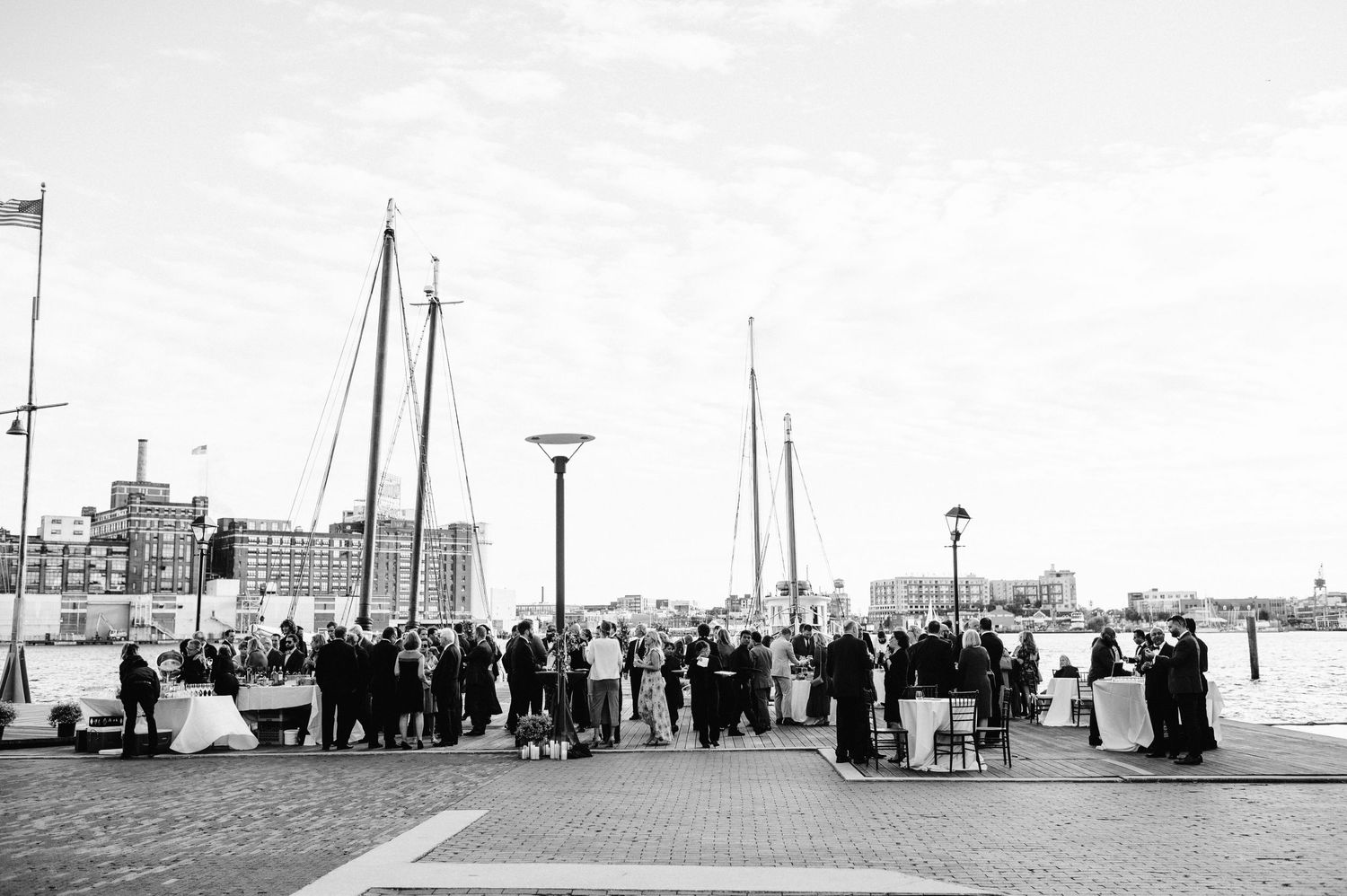 A large group gathers on a waterfront promenade with sailboat masts visible in the background.
