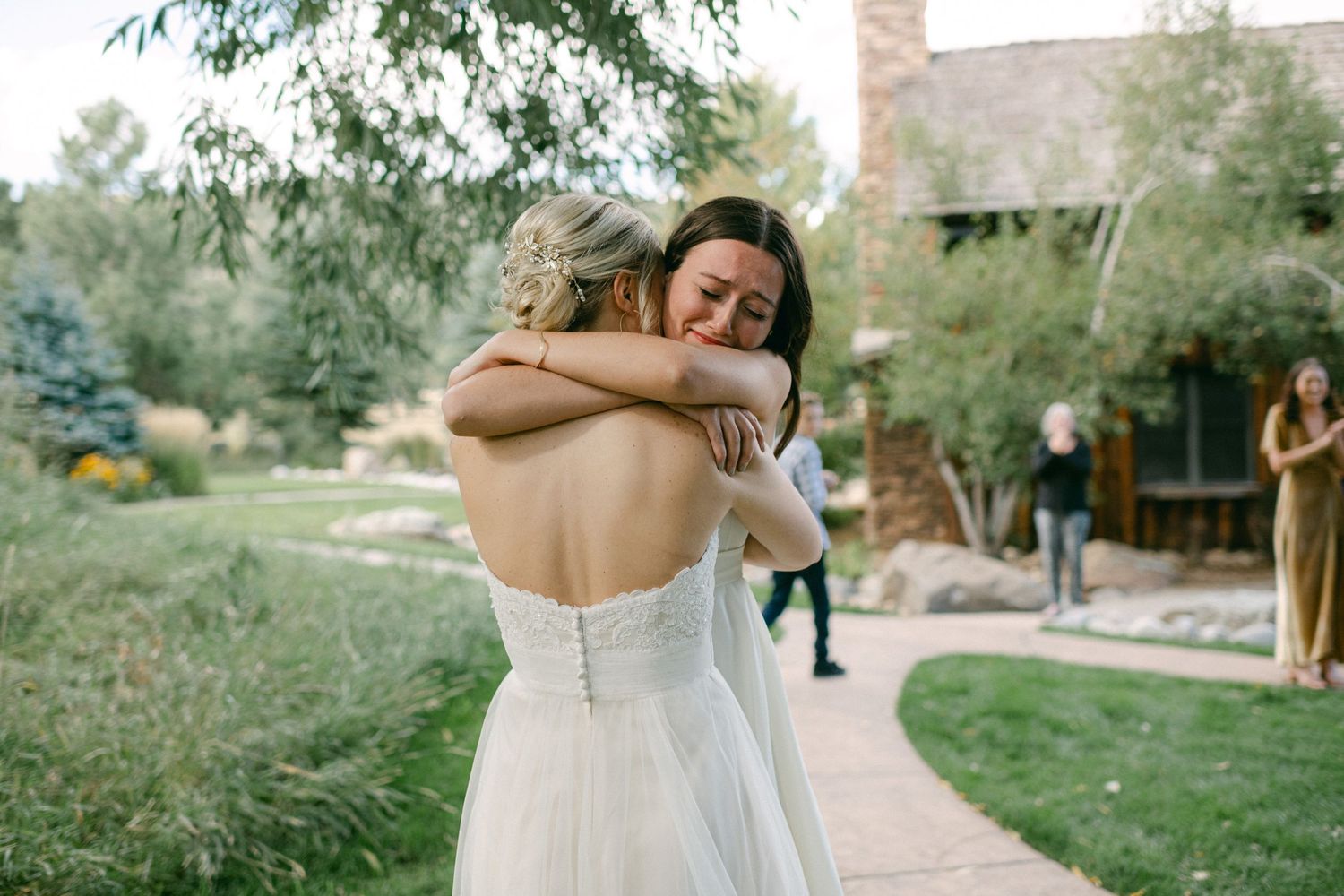 Two people in white dresses sharing an emotional embrace during an outdoor wedding celebration.