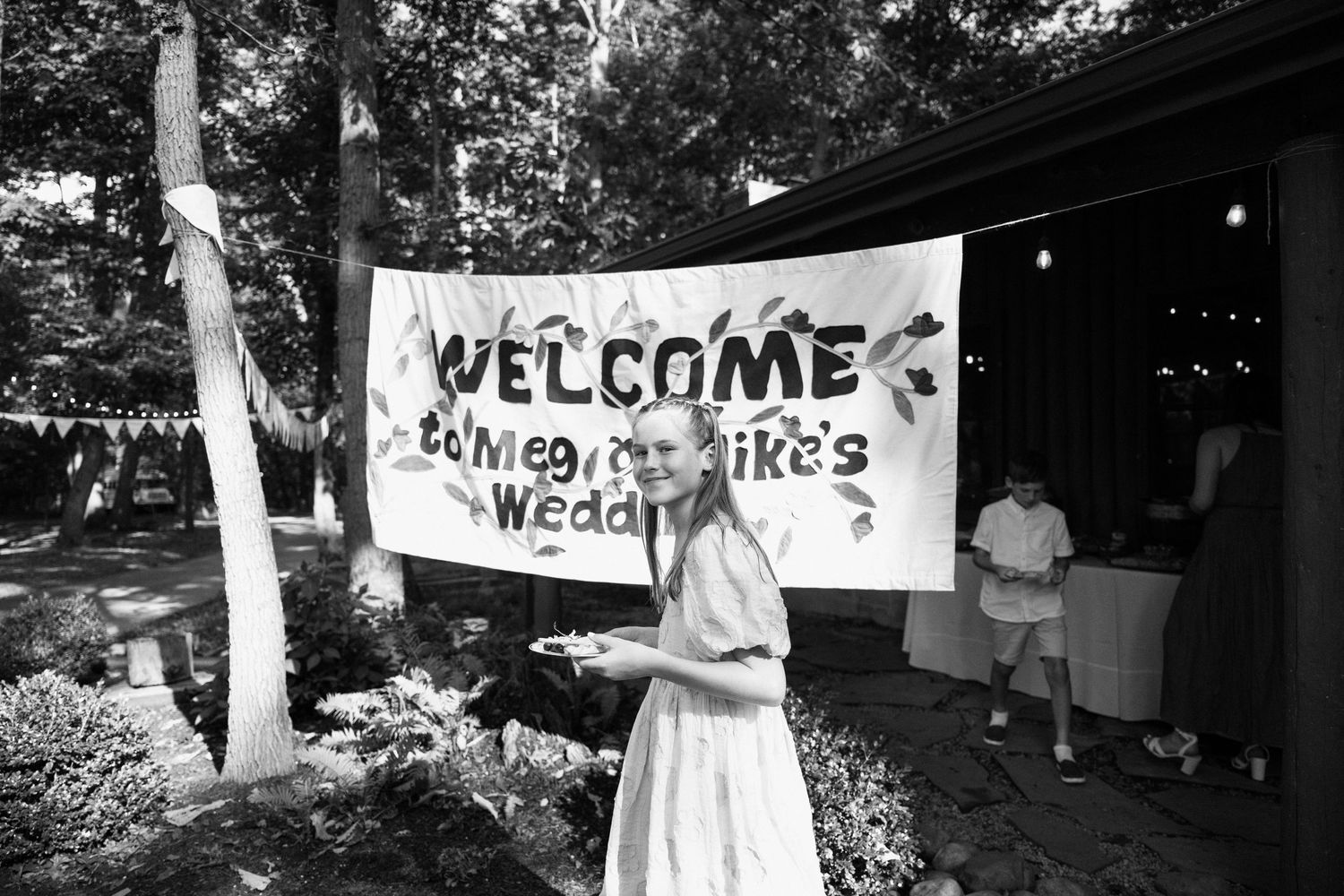 A welcome sign hangs at an outdoor wedding reception surrounded by trees.
