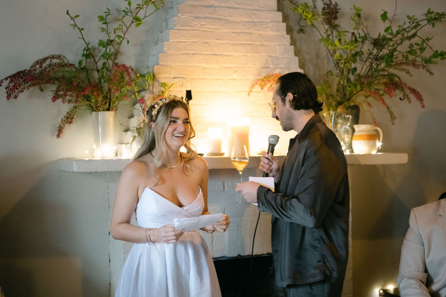 Bride in white dress and groom sharing a lighthearted conversation against a candlelit backdrop with greenery.