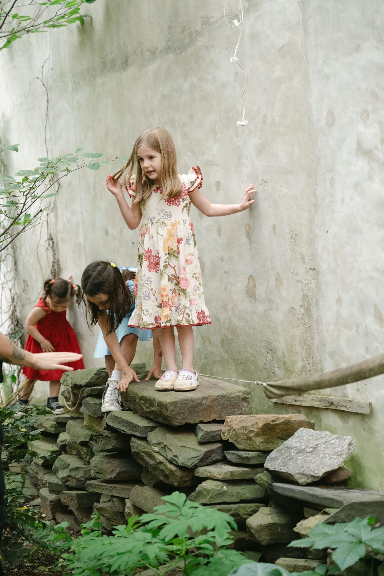 Two children playing on a stone wall near a garden beside a weathered gray concrete wall while wearing summer dresses.