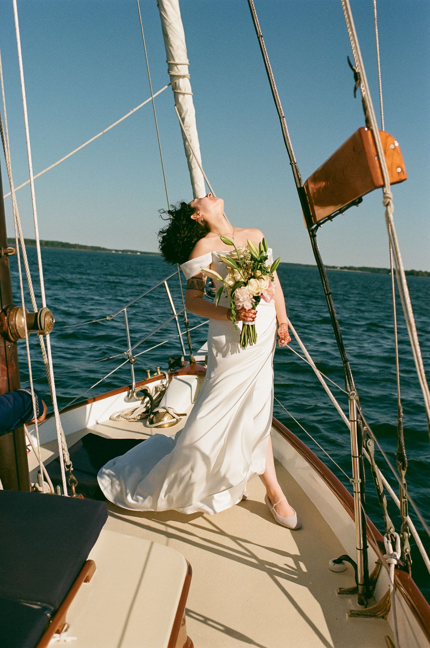 Bride in white gown holding bouquet poses on sailboat deck with blue ocean backdrop on sunny day.