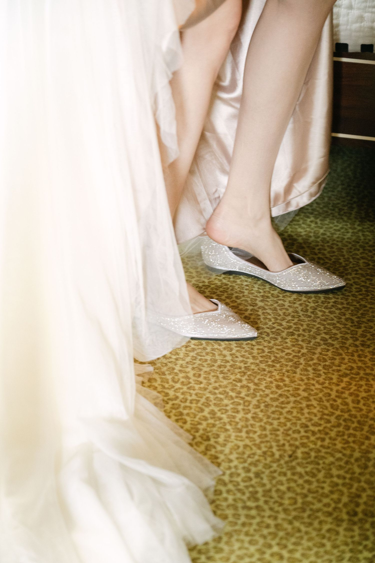 Bride wearing sparkling silver flat wedding shoes beneath an elegant white gown on a leopard print carpet.
