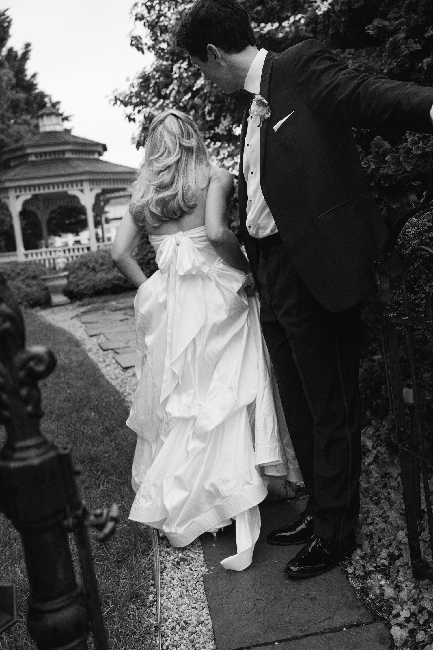 Black and white photo of bride in ruffled gown and groom in tuxedo sharing intimate moment by garden gazebo.