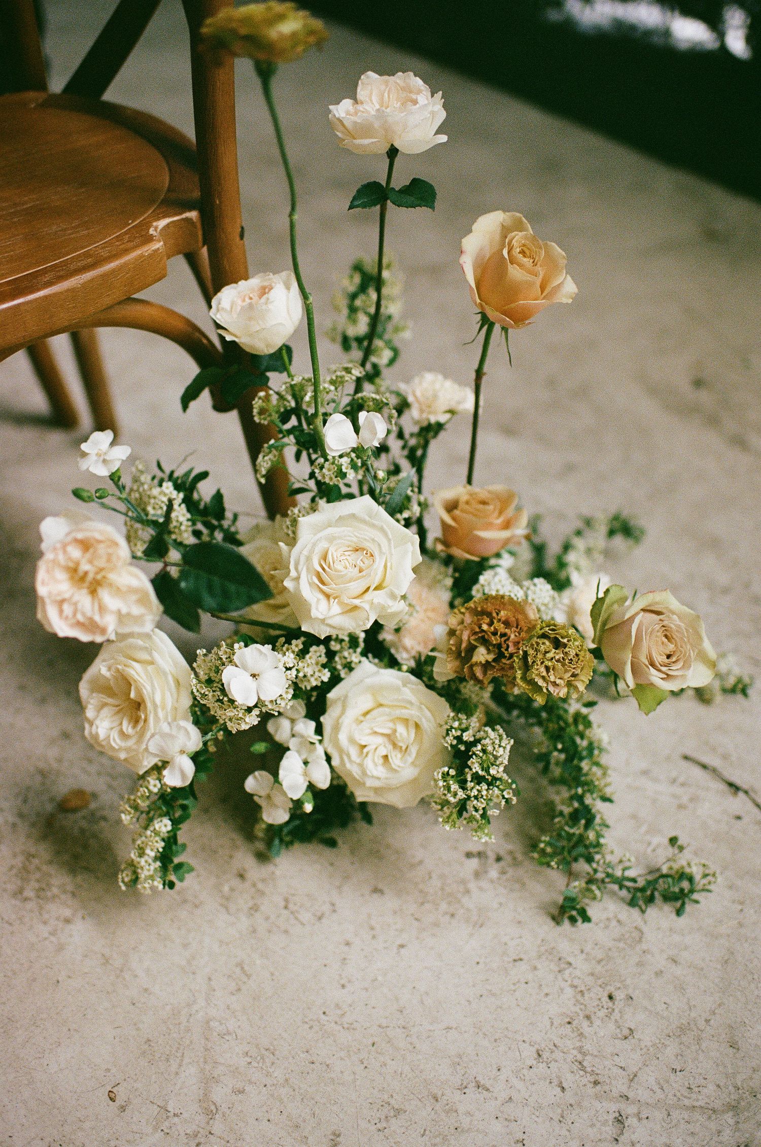 Elegant floor arrangement of cream and peach roses with greenery beside a wooden chair.