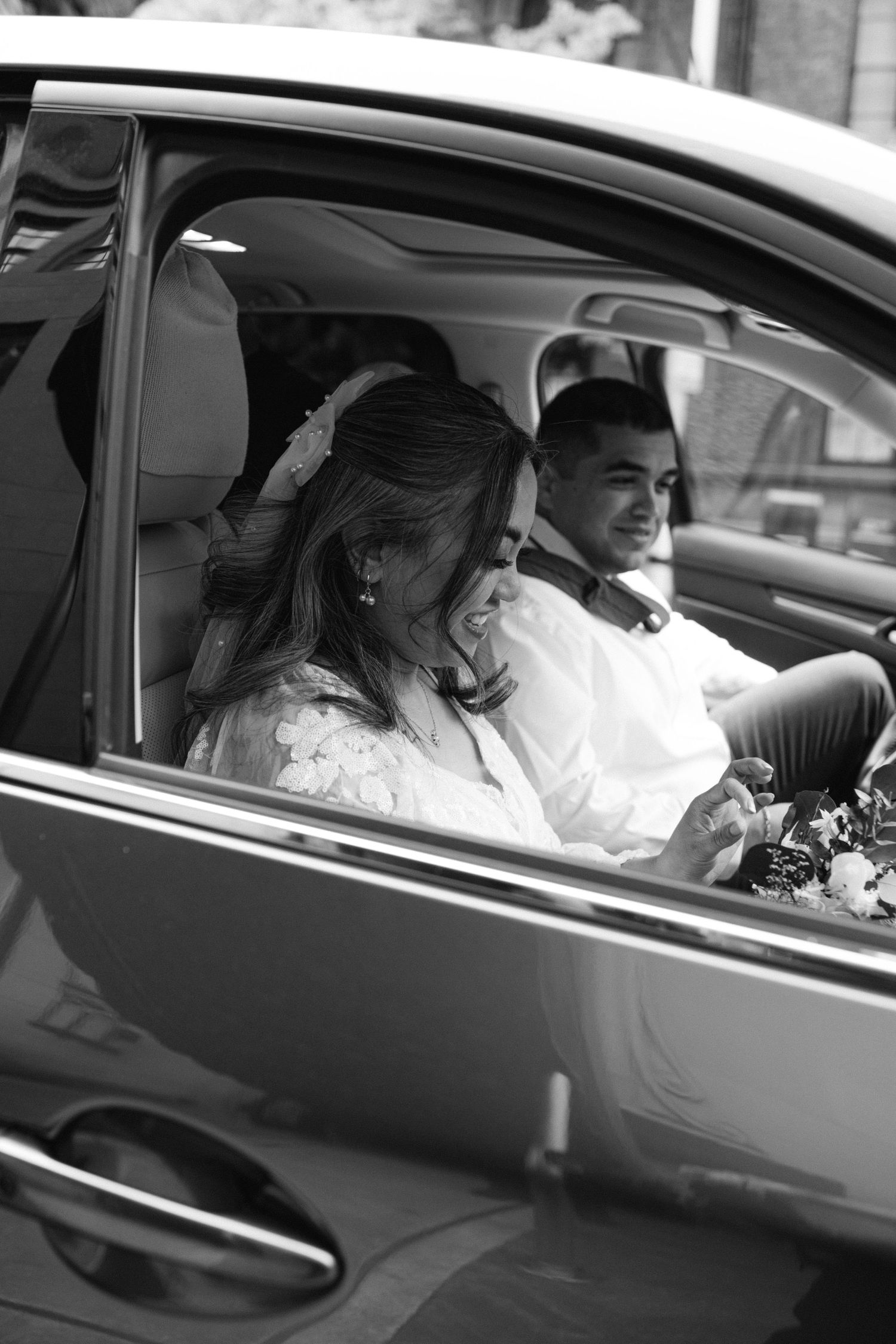 A couple sits in a car together with flowers visible through the window.