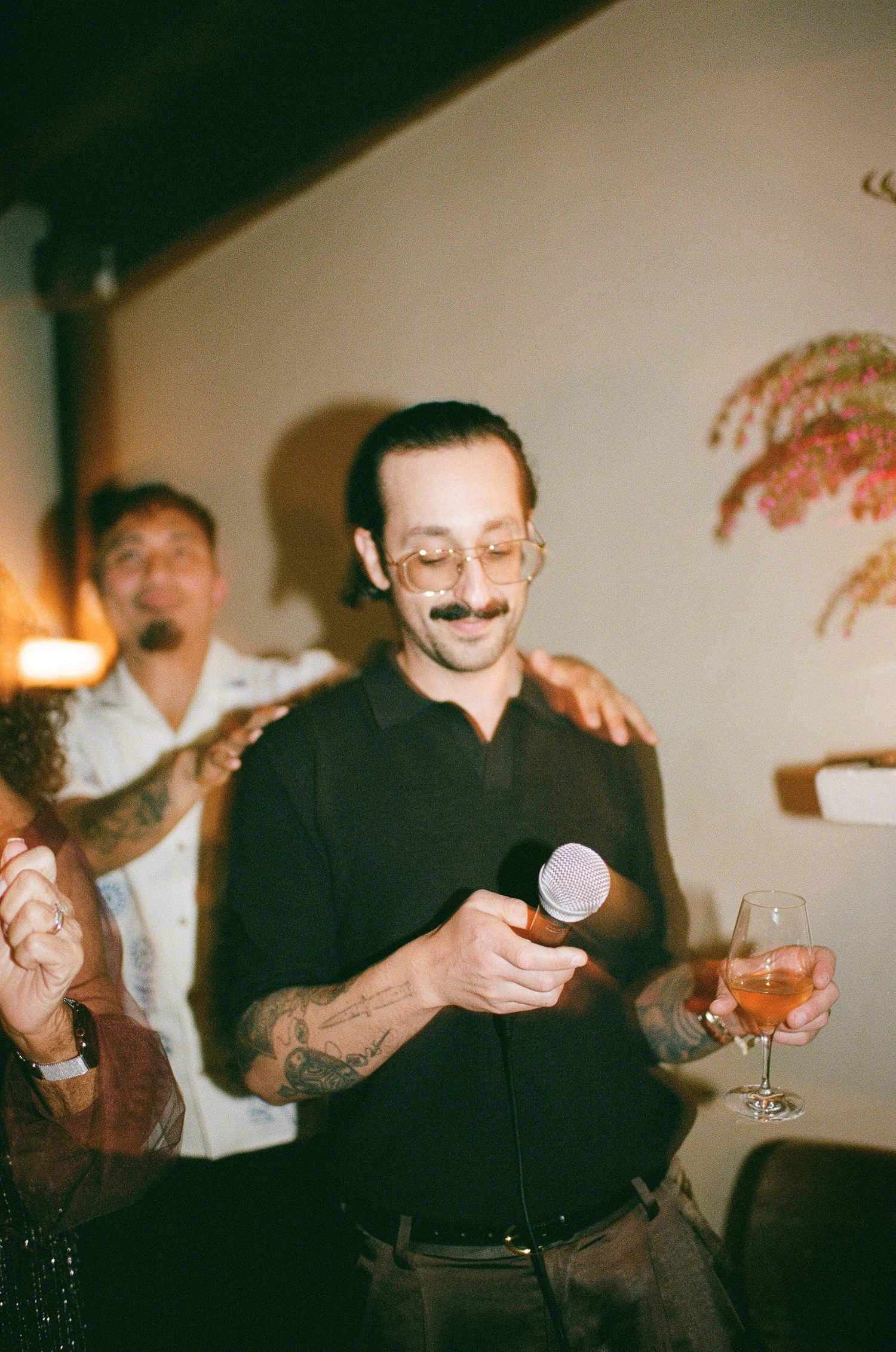 Person in black shirt smiling while holding microphone and cocktail at indoor party with decorative wall art.