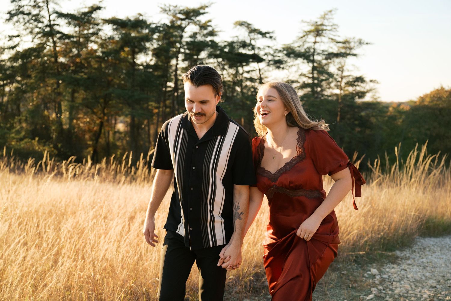 Two people walk hand in hand through a golden field at sunset with tall trees in the background.