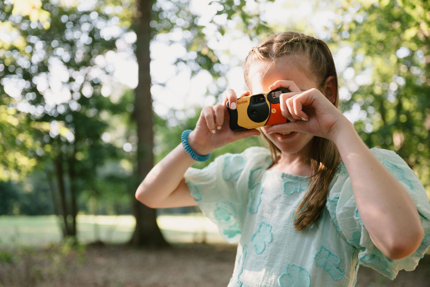 Young child in light blue dress looking through a toy camera outdoors.