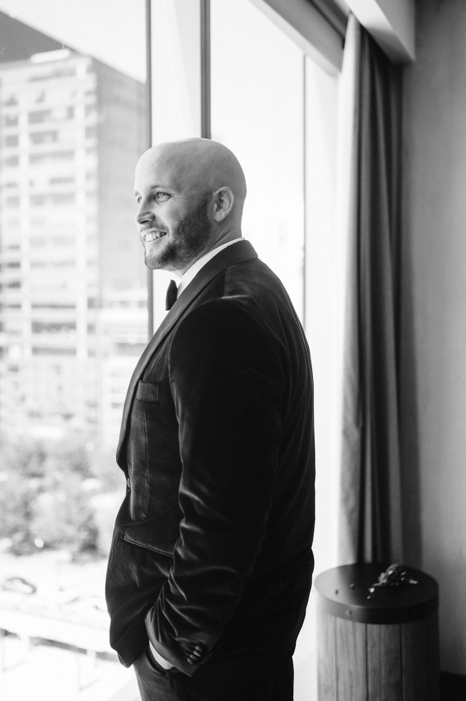 A black and white portrait of a groom smiling while looking out a window.