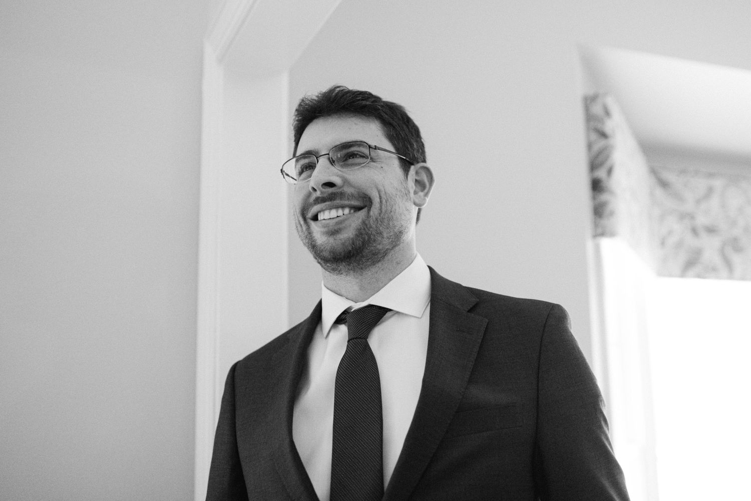 A portrait of a groom wearing glasses and a dark suit laughing joyfully on his wedding day.