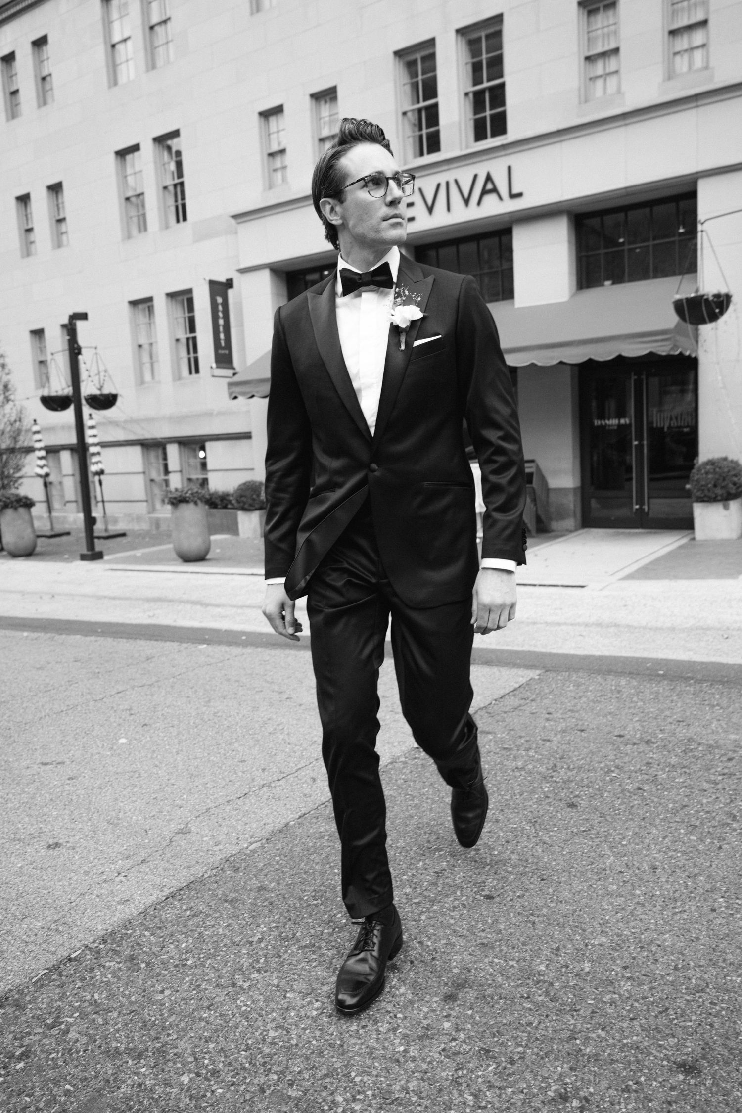 A formal-dressed man in a black tuxedo and glasses walks confidently past an urban building.