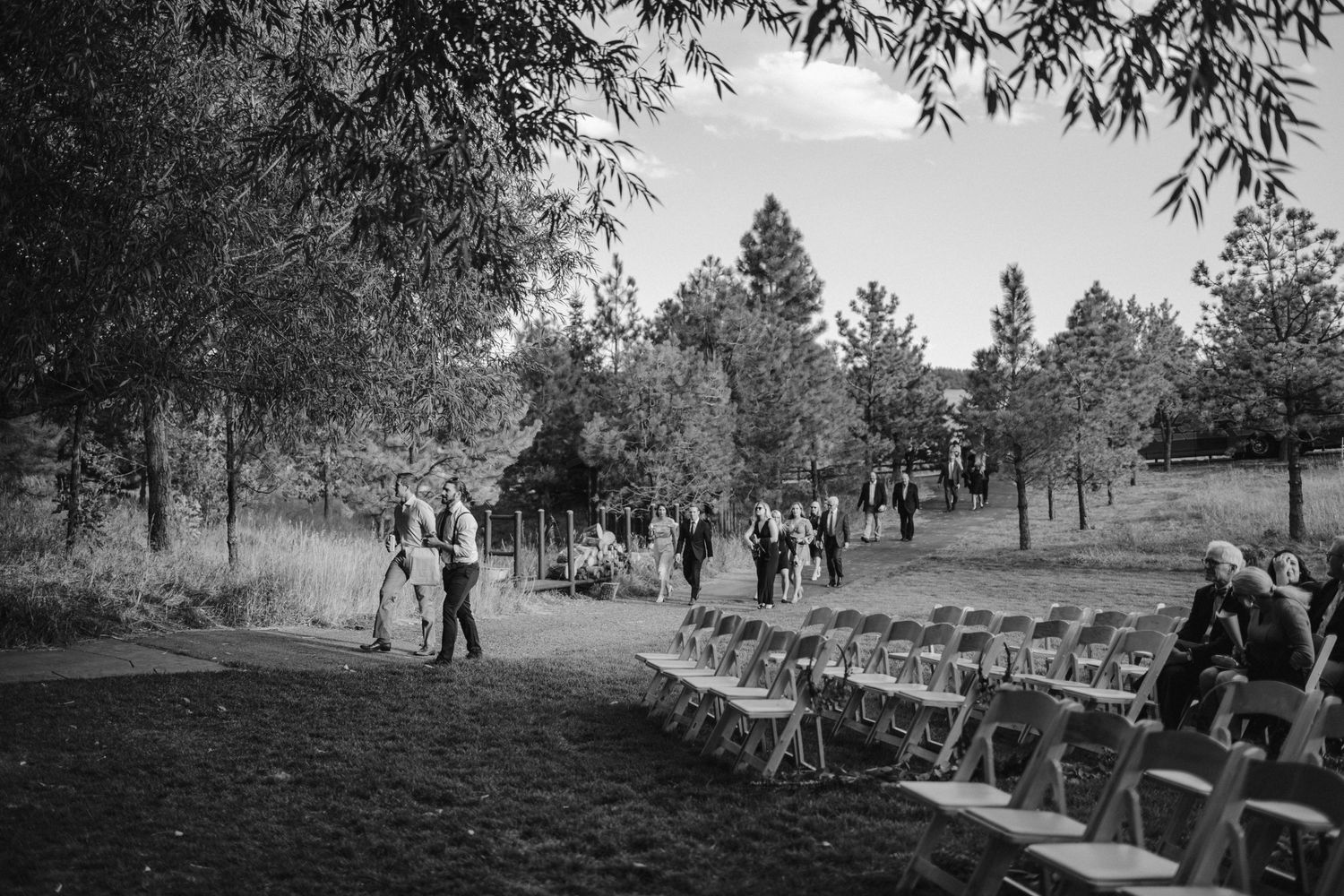 Black and white photo of an outdoor wedding ceremony setup with rows of chairs and guests in a wooded setting.