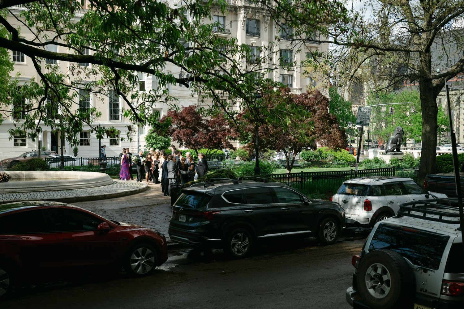 Urban street view with cars parked beneath blooming spring trees and rows of white apartment buildings.