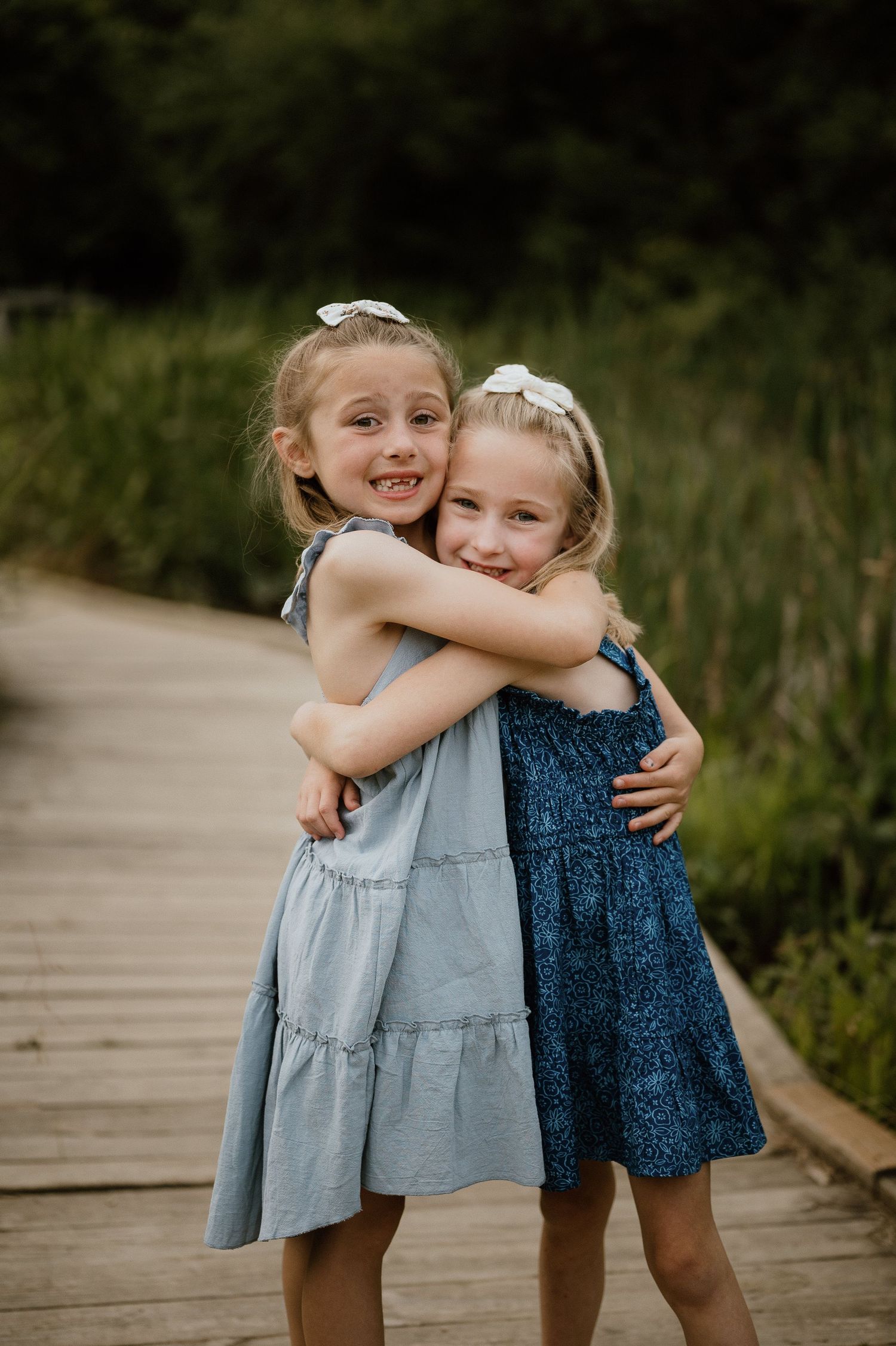 Two children in blue dresses share a warm embrace while standing on a wooden walkway.