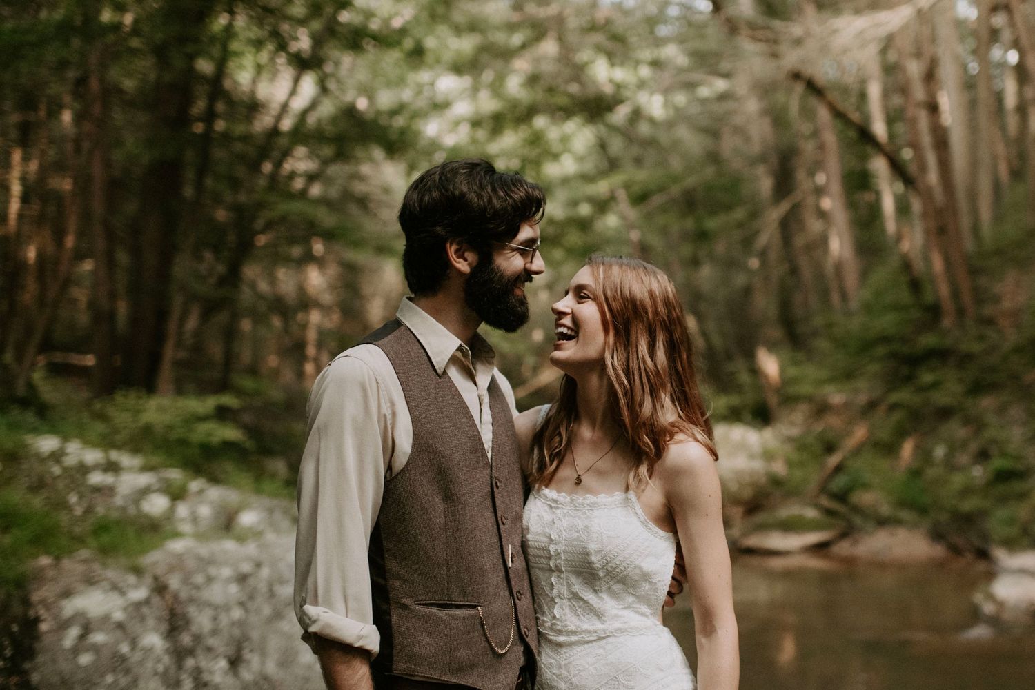 A romantic moment between newlyweds in a scenic forest setting with a stream backdrop.